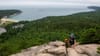 A hiker ascends the rocky Beehive Loop Trail in Acadia National Park, with views overlooking Sand Beach and the Atlantic Ocean.